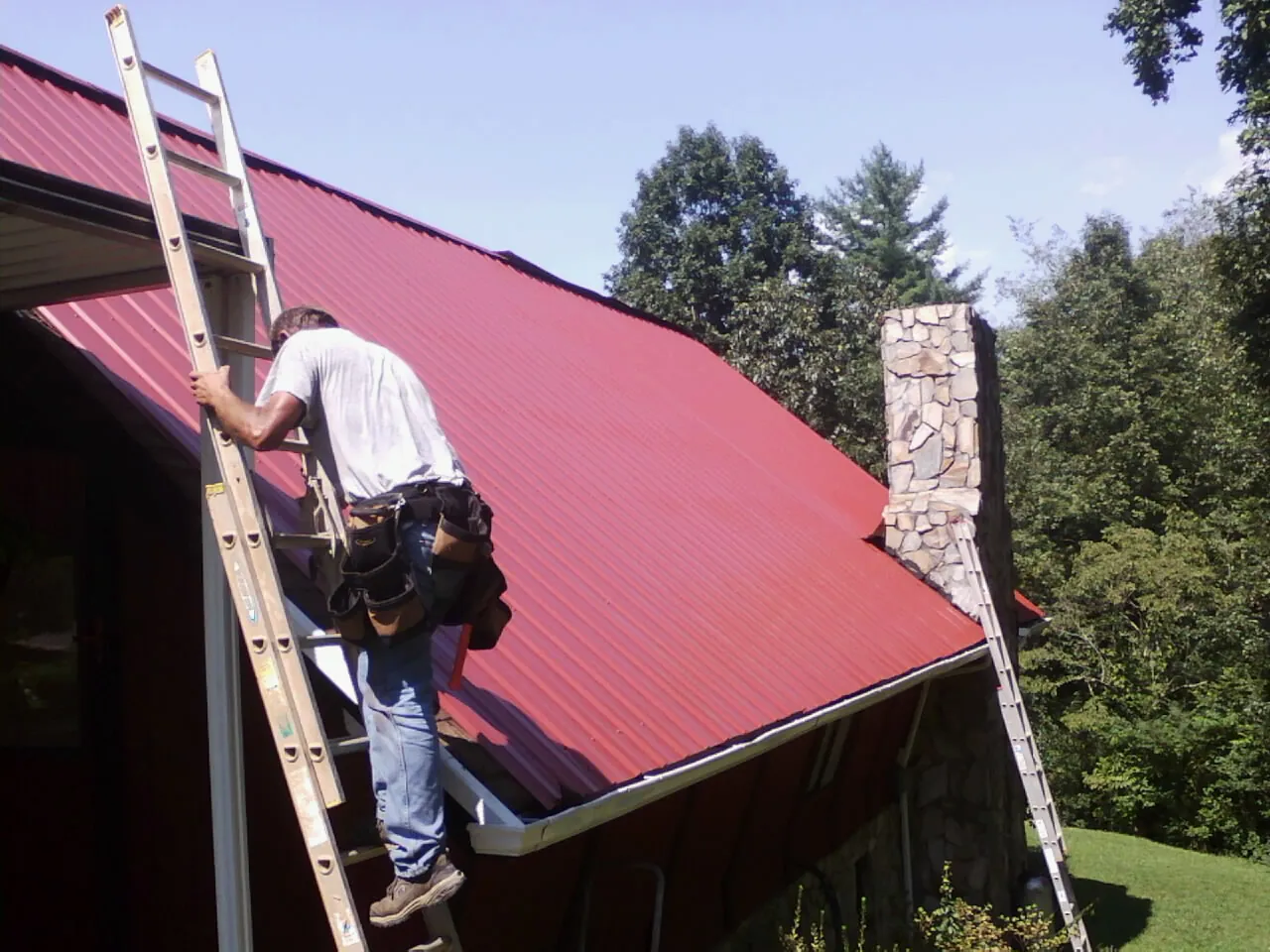 Roofer on a ladder beside a red metal roof and stone chimney — Soffit & Fascia Repair by Roofing Brothers in Guilford